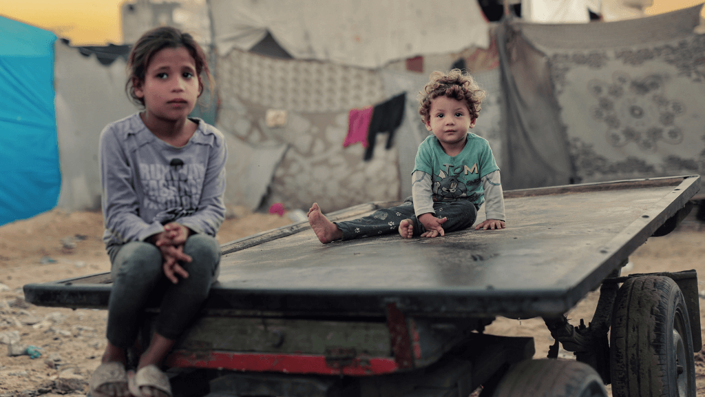 Two children sitting on a cart in an outdoor setting, surrounded by makeshift structures.