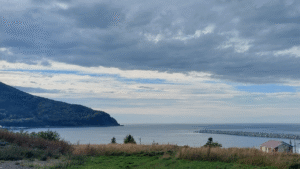 Coastal view with cloudy sky, green landscape, and distant mountain by the ocean.