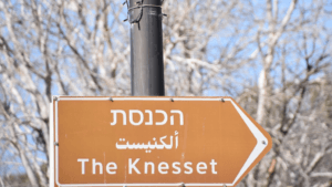 Street sign pointing to The Knesset in Hebrew, Arabic, and English on a clear day with trees in the background.